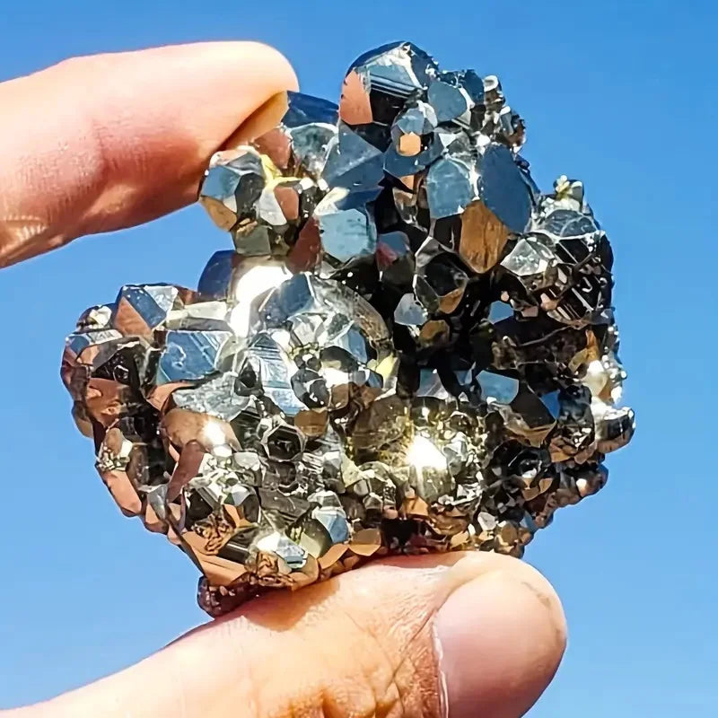 Natural pyrite rough stone held in hand against a blue sky, showcasing its shiny, irregular surface and unique crystal formations.